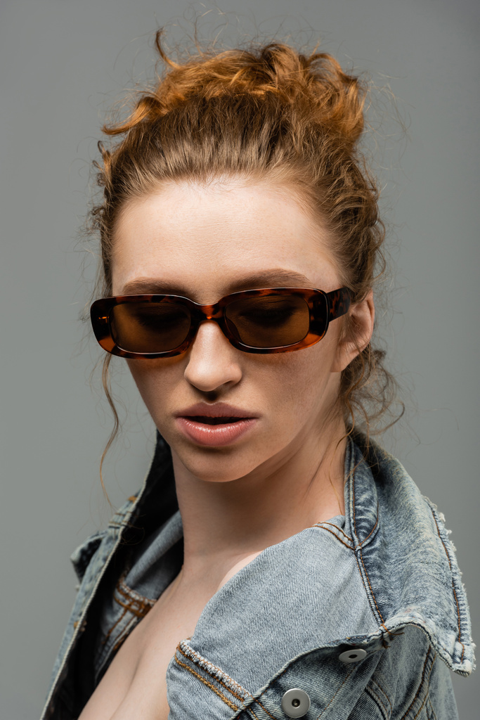 Κομψή και με αυτοπεποίθηση νεαρή κοκκινομάλλα μοντέλο με φυσικό μακιγιάζ σε γυαλιά ηλίου ποζάρουν σε τζιν σακάκι και στέκεται απομονωμένη σε γκρι φόντο, μοντέρνα έννοια αντηλιακής προστασίας, μοντέλο μόδας  - Φωτογραφία, εικόνα