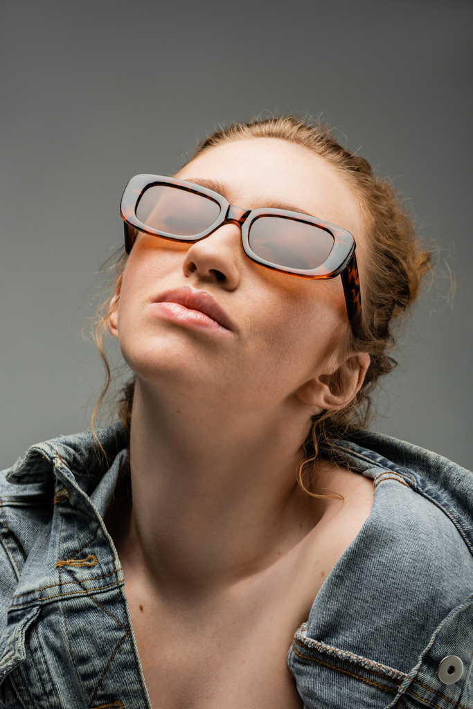 Porträt einer rothaarigen und sommersprossigen jungen Frau mit stylischer Sonnenbrille und Jeansjacke, die unter Beleuchtung isoliert auf grauem Hintergrund steht, trendiges Sonnenschutzkonzept, Modemodel  - Foto, Bild