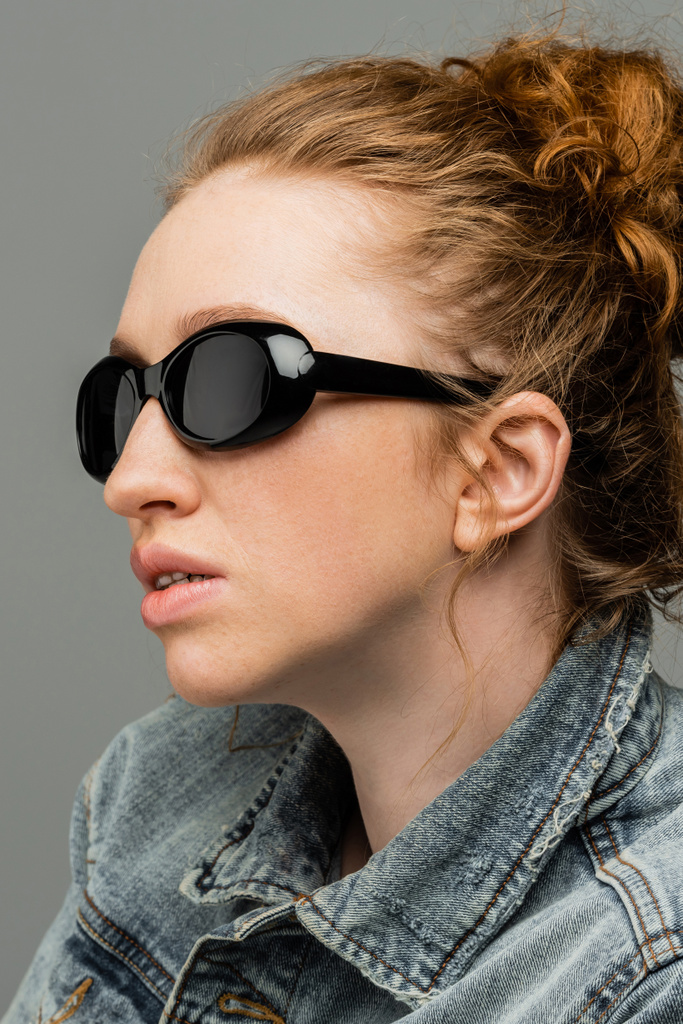 Portrait of freckled and red haired woman with natural makeup wearing stylish sunglasses and denim jacket isolated on grey background, trendy sun protection concept, fashion model  - Photo, Image