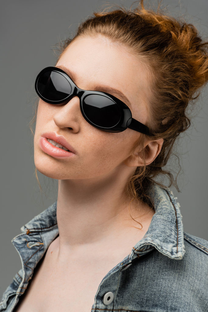 Portrait of young redhead and freckled woman in fashionable sunglasses and denim jacket standing isolated on grey background, trendy sun protection concept, fashion model  - Photo, Image