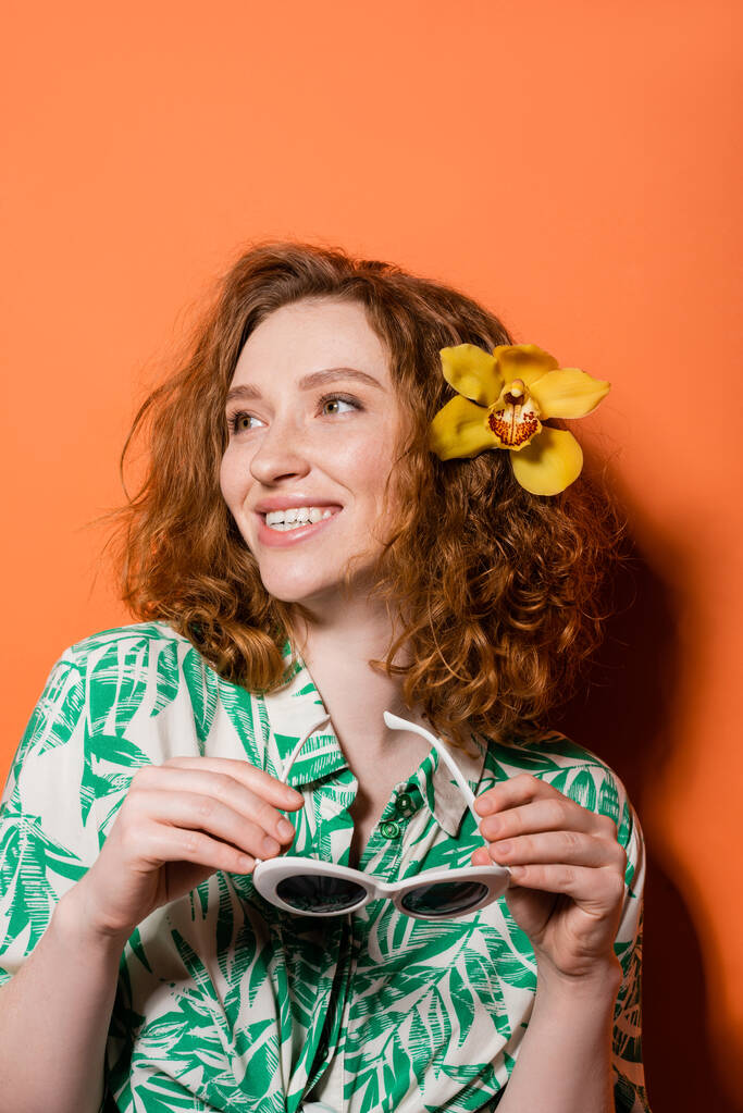 Giovane donna rossa sorridente con fiore di orchidea in capelli che indossa camicetta con stampa floreale e tenendo gli occhiali da sole mentre in piedi su sfondo arancione, estate casual e concetto di moda, Cultura giovanile - Foto, immagini