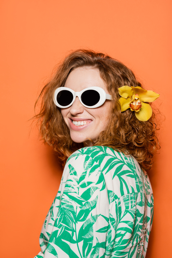 Jovem mulher positiva com cabelo vermelho e flor de orquídea posando em óculos de sol e blusa com estampa floral em fundo laranja, verão casual e conceito de moda, Cultura Jovem - Foto, Imagem