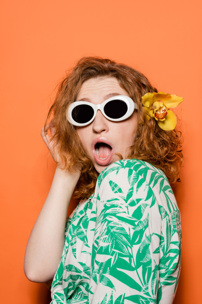 Шокированная молодая рыжая женщина с орхидеей цветок и солнцезащитные очки, позируя в блузке с цветочным узором и стоя на оранжевом фоне, летом случайные и моды концепции, молодежная культура - Фото, изображение