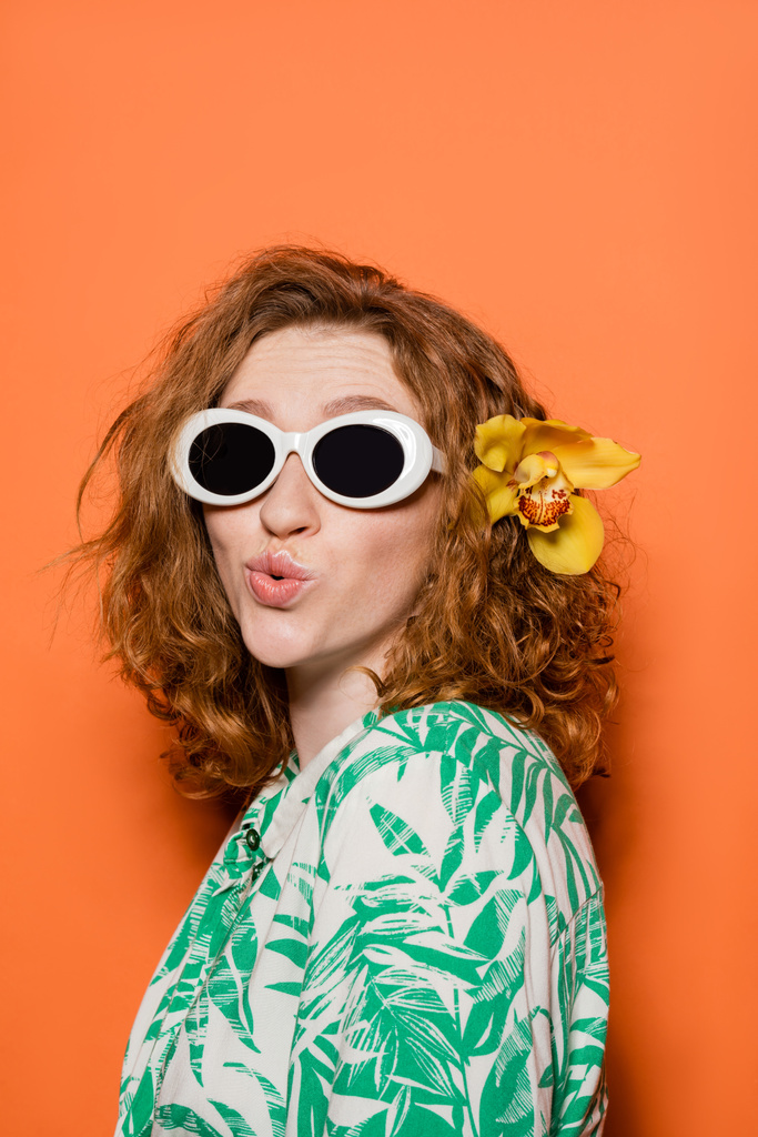 Młoda kobieta z czerwonymi włosami i orchideą kwiat noszenia okularów przeciwsłonecznych i bluzki z kwiatowym wzorem podczas dąsania warg i stojąc na pomarańczowym tle, lato casual i koncepcja mody, kultura młodzieży - Zdjęcie, obraz