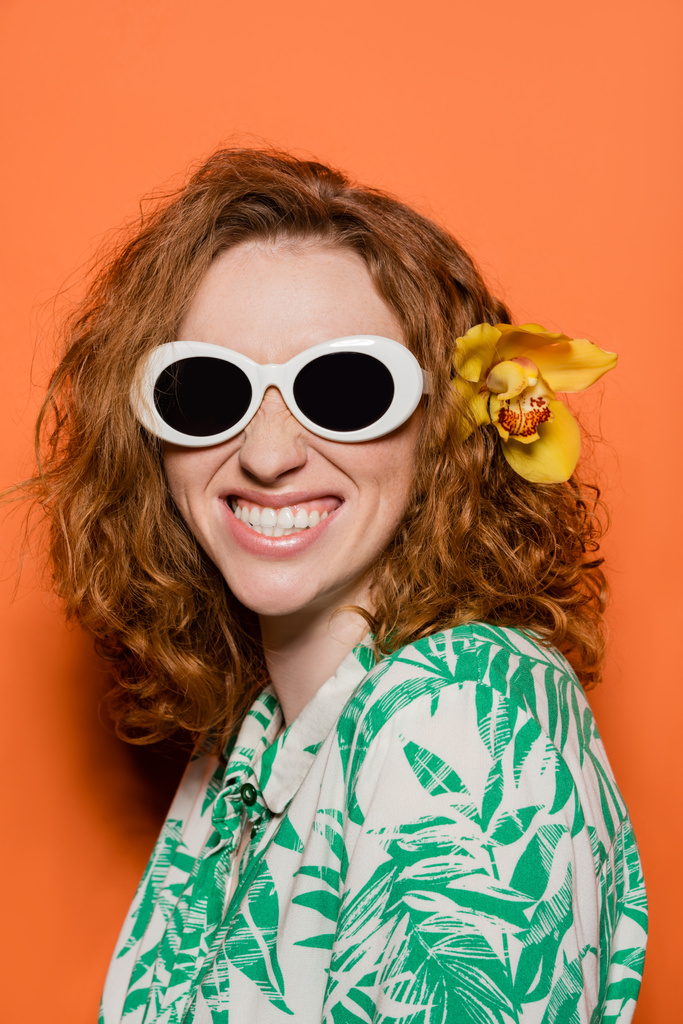Позитивная женщина с цветком орхидеи в рыжих волосах в солнцезащитных очках и блузке с цветочным узором во время позирования и стоя на оранжевом фоне, летняя повседневная и модная концепция, молодежная культура - Фото, изображение