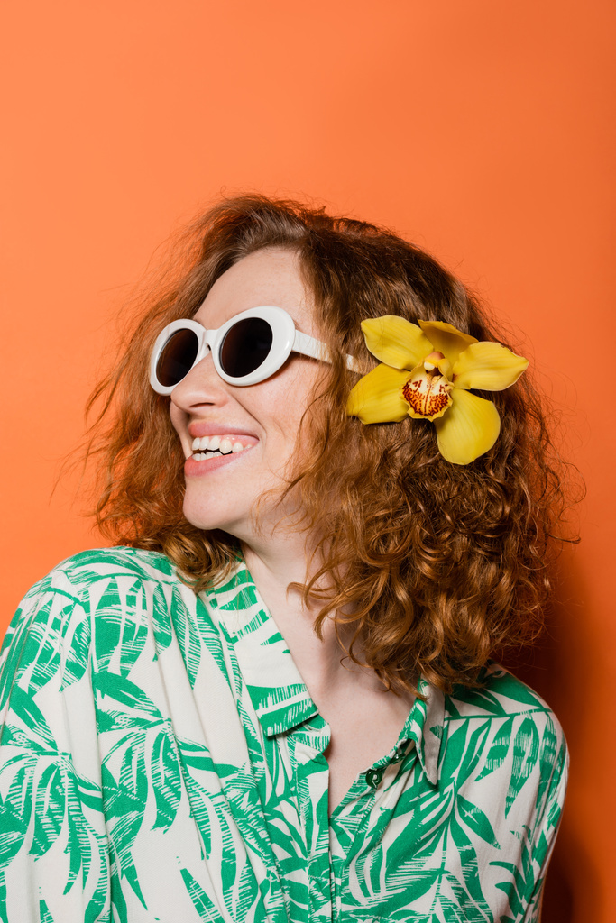 Щаслива і впевнена молода руда жінка з орхідеями в волоссі, стильні сонячні окуляри і сучасна блузка стоять на помаранчевому тлі, літні випадкові і модні концепції, Молодіжна культура - Фото, зображення