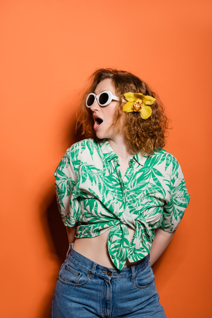 Шокированная рыжая женщина в солнцезащитных очках и цветке орхидеи в блузке с цветочным принтом и джинсах, позируя и стоя на оранжевом фоне, летняя повседневная и модная концепция, молодежная культура - Фото, изображение