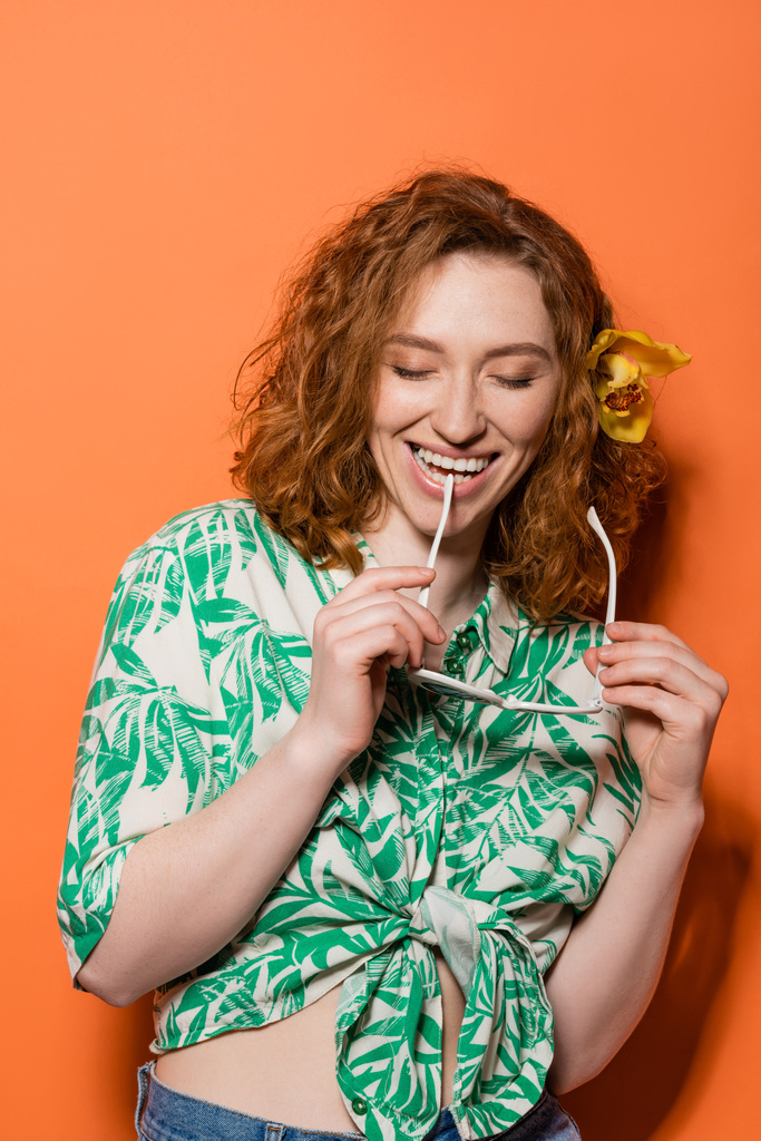 Positive redhead woman with orchid flower holding fashionable sunglasses and closing eyes while standing on orange background, summer casual and fashion concept, Youth Culture - Photo, Image