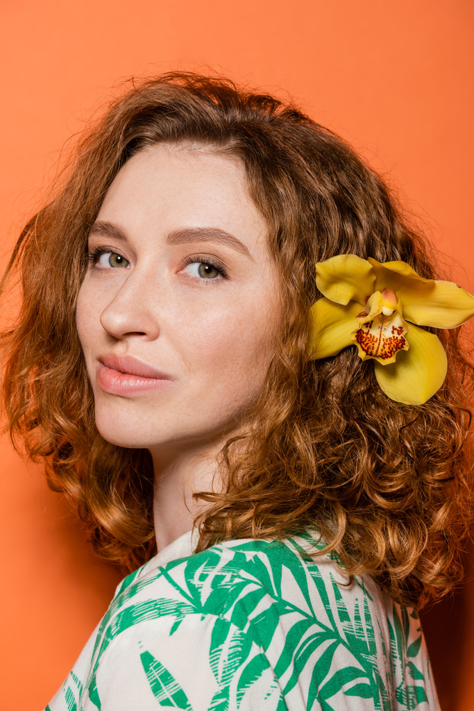 Πορτρέτο της νεαρής κοκκινομάλλας με ορχιδέα λουλούδι στα μαλλιά και φυσικό μακιγιάζ κοιτάζοντας κάμερα, ενώ στέκεται σε πορτοκαλί φόντο, καλοκαίρι casual και μόδα έννοια, Νεολαία Πολιτισμού - Φωτογραφία, εικόνα