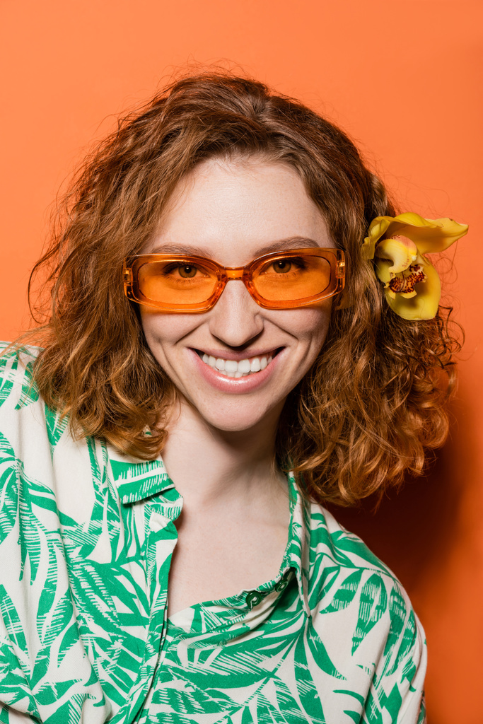 Porträt einer lächelnden und stilvollen jungen Frau mit Orchideenblüte im roten Haar, die in die Kamera blickt und mit Sonnenbrille auf orangefarbenem Hintergrund posiert, Sommer- und Modekonzept, Jugendkultur - Foto, Bild