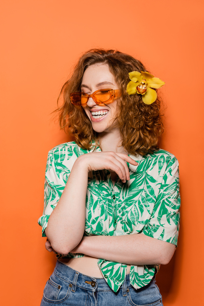 Happy young redhead woman with orchid flower and sunglasses posing in blouse with floral print and jeans on orange background, summer casual and fashion concept, Youth Culture - Photo, Image
