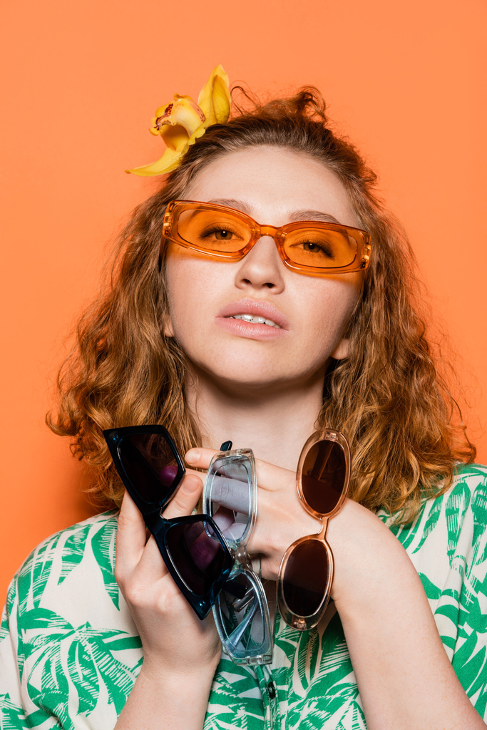 Portrait of stylish young woman with orchid flower in red hair holding sunglasses and looking at camera while standing on orange background, summer casual and fashion concept, Youth Culture - Photo, Image