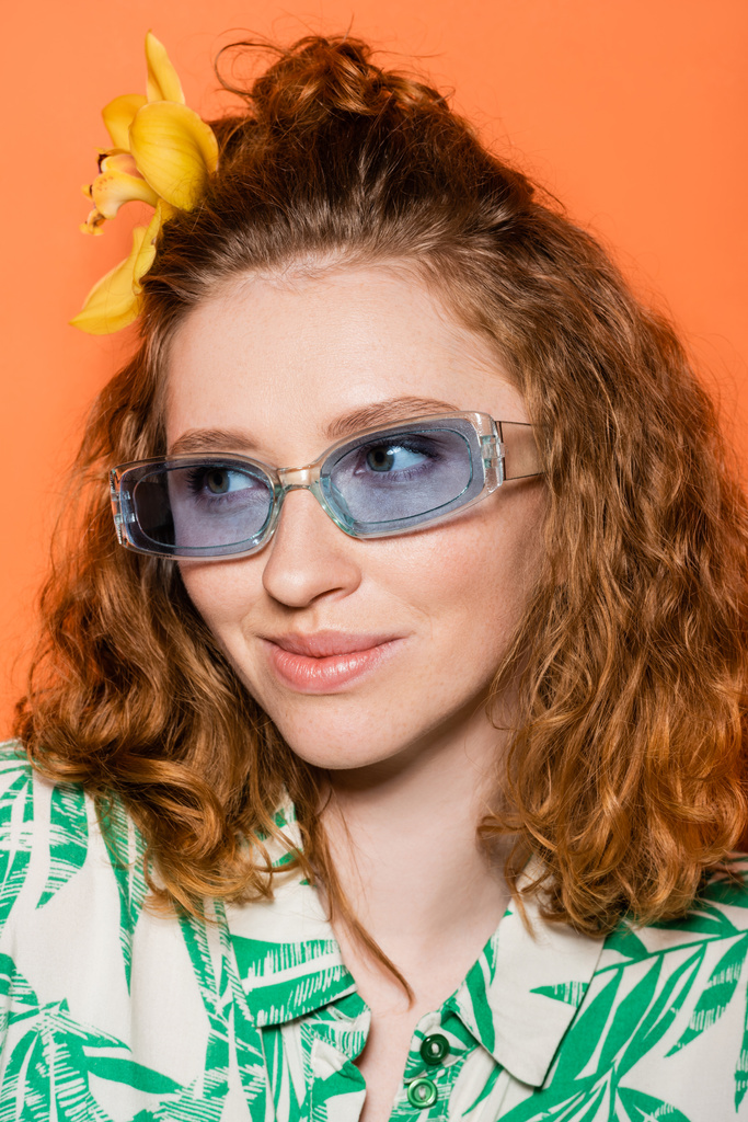 Retrato de modelo jovem ruiva com flor de orquídea no cabelo usando óculos de sol azuis e blusa na moda com padrão floral isolado em laranja, roupa casual elegante e conceito de vibrações de verão - Foto, Imagem