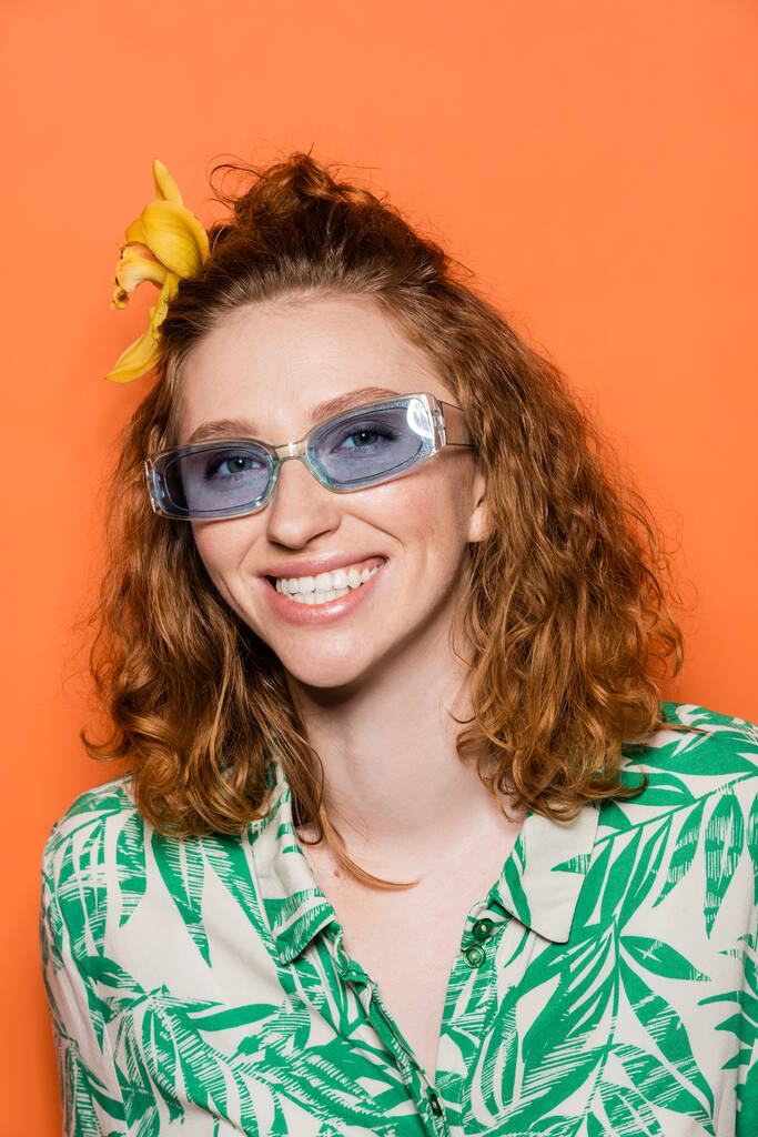 Iloinen nuori punapää nainen, jolla on orkidea kukka hiuksissa ja siniset aurinkolasit katselevat kameraa seistessään eristettynä oranssiin, tyylikkääseen rentoon asuun ja kesätunnelmaan. - Valokuva, kuva