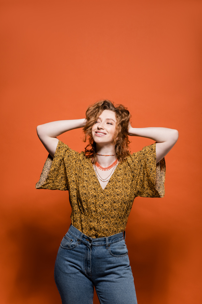 Mujer pelirroja joven de moda en blusa con impresión abstracta moderna y jeans tocando la cabeza sobre fondo naranja, elegante atuendo casual y concepto de vibraciones de verano, Cultura Juvenil - Foto, imagen