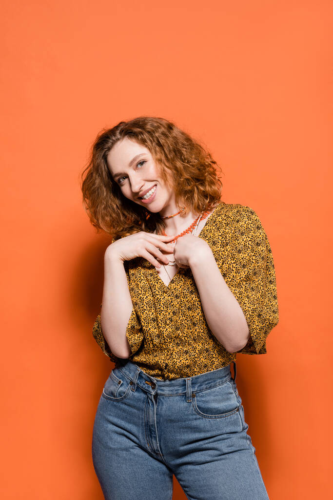 Joyful and stylish young redhead woman in blouse with abstract pattern and jeans touching necklaces and standing on orange background, stylish casual outfit and summer vibes concept, Youth Culture - Photo, Image
