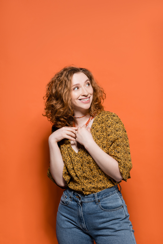 Cheerful and stylish young redhead woman in casual modern blouse and jeans touching necklaces and standing on orange background, stylish casual outfit and summer vibes concept, Youth Culture - Photo, Image