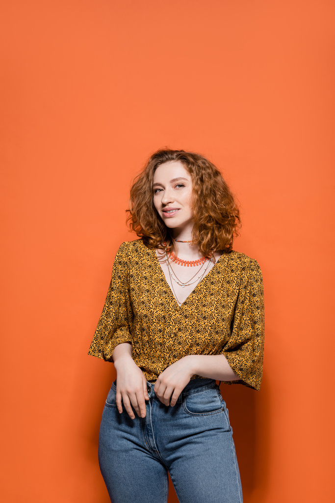 Modny młody rudy model w żółtej bluzce z abstrakcyjnym wzorem i dżinsami uśmiechniętymi do kamery i stojącymi na pomarańczowym tle, stylowy strój casual i letnia koncepcja wibracji, kultura młodzieży - Zdjęcie, obraz
