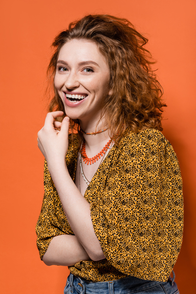 Portrait de jeune femme rousse en chemisier avec imprimé abstrait et jeans riant et regardant la caméra sur fond orange, tenue décontractée élégante et concept d'ambiance estivale, Culture de la jeunesse - Photo, image