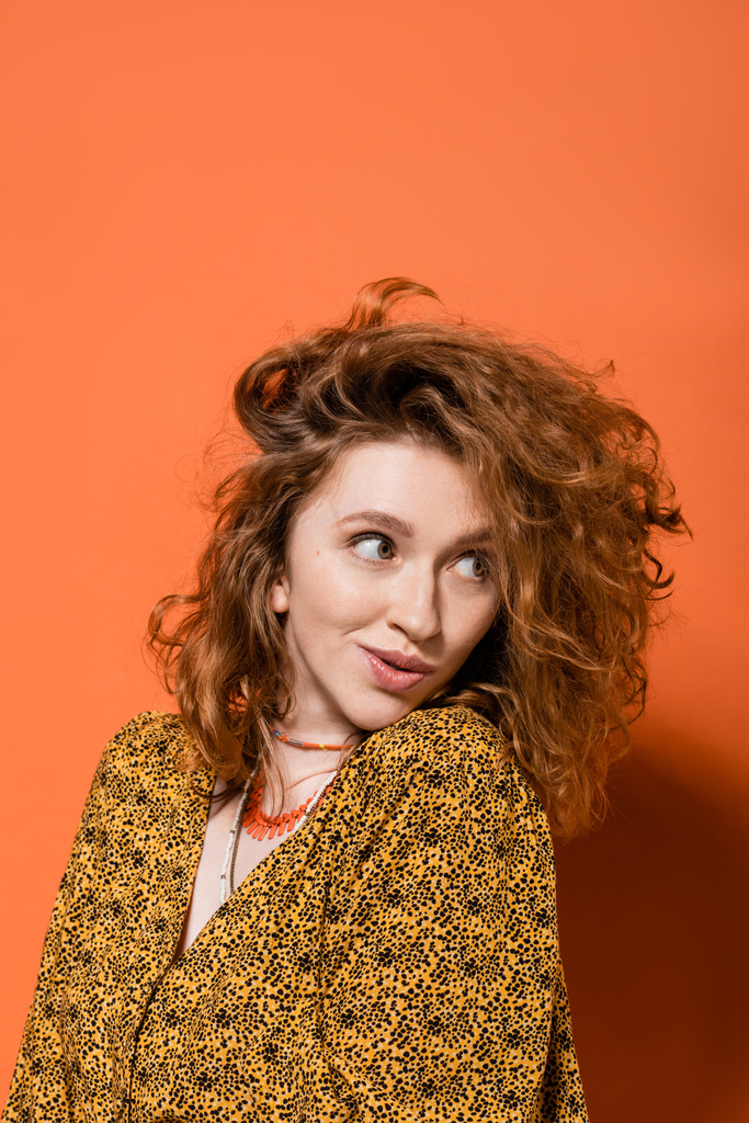 Astonished young red haired woman in yellow blouse with abstract pattern and necklaces looking away and standing on orange background, stylish casual outfit and summer vibes concept, Youth Culture - Photo, Image