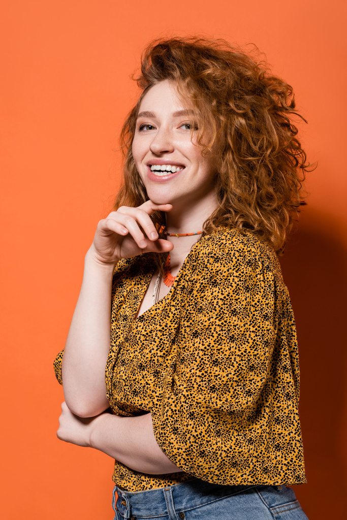 Boldog fiatal vörös hajú nő nyakláncban, sárga blúz elvont mintával, ahogy a kamerába néz és narancssárga háttérrel pózol, stílusos alkalmi ruha és nyári vibrálás koncepció, Ifjúsági Kultúra - Fotó, kép