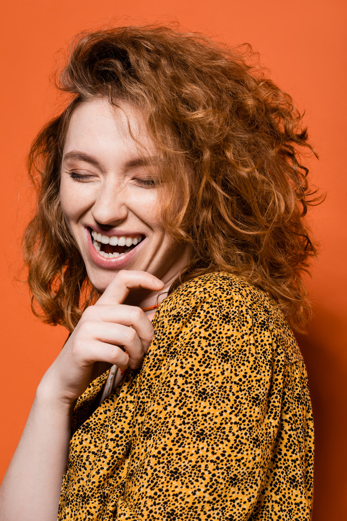 Portret van jonge en positieve roodharige vrouw in gele blouse met abstract patroon met gesloten ogen op oranje achtergrond, stijlvolle casual outfit en zomer vibes concept, Jeugdcultuur - Foto, afbeelding