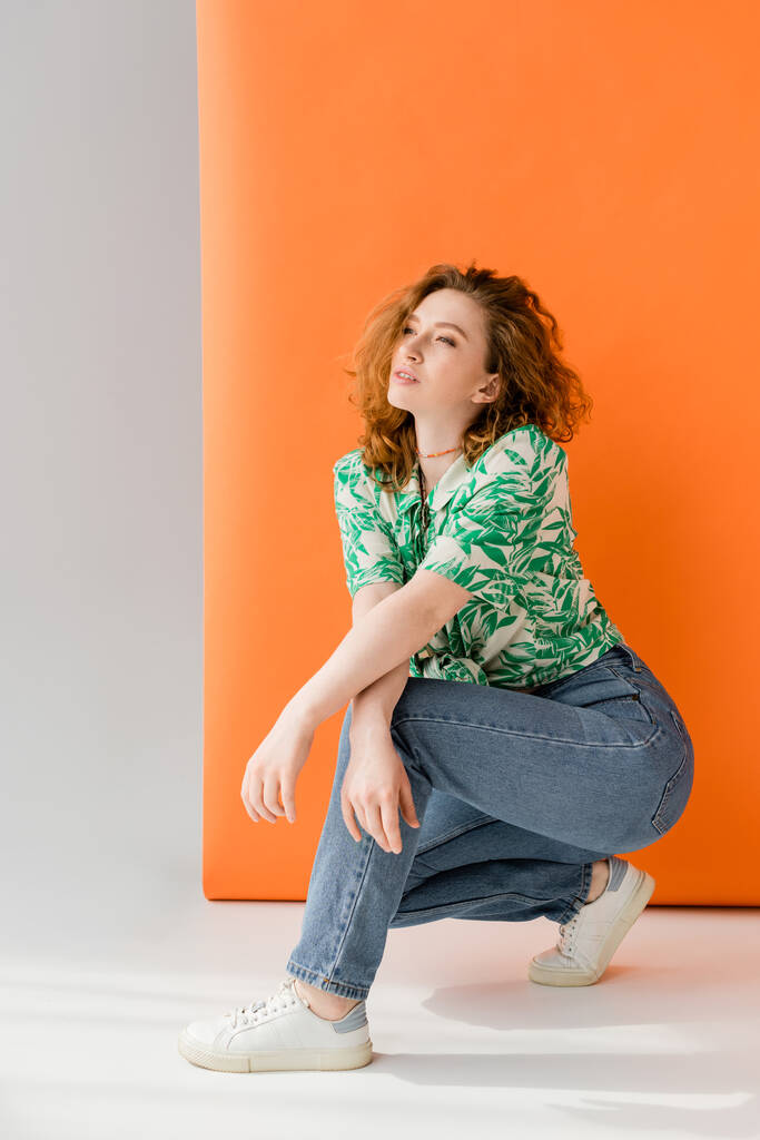 Full length of young red haired woman in blouse with floral pattern and jeans looking away while posing on orange and grey background, trendy casual summer outfit concept, Youth Culture - Photo, Image