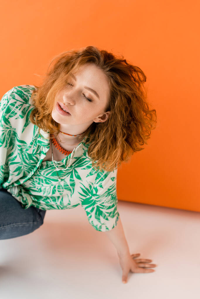 Stylish young redhead woman in blouse with floral pattern, necklaces and jeans posing with closed eyes on orange background, trendy casual summer outfit concept, Youth Culture - Photo, Image