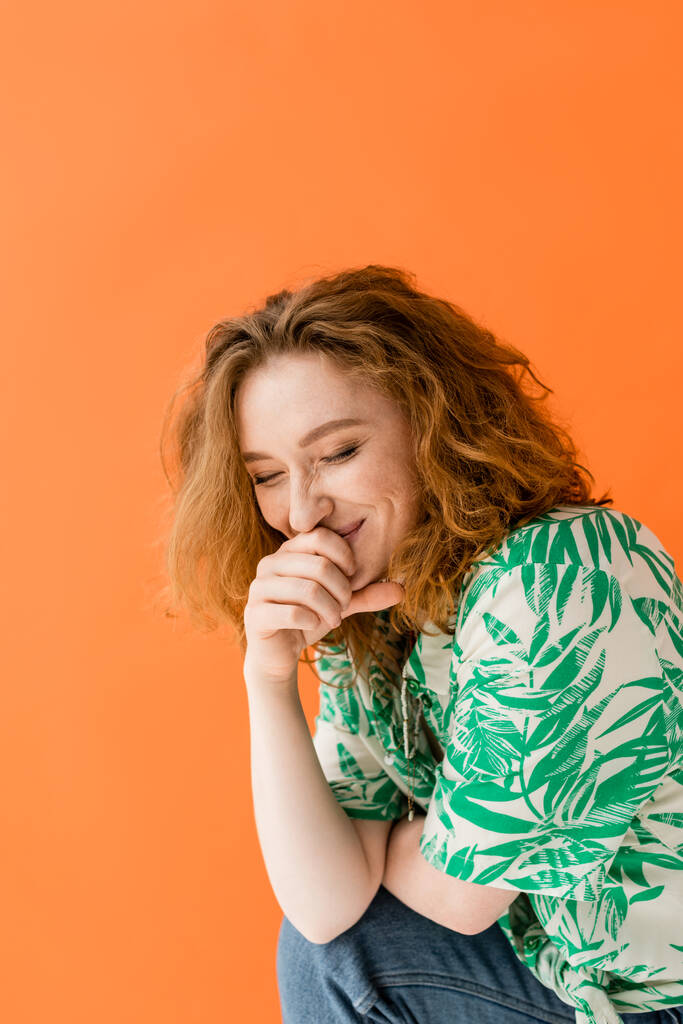 Jovem mulher ruiva alegre com olhos fechados em blusa com padrão floral e jeans rindo enquanto posando isolado no conceito de roupa de verão casual laranja, na moda, Cultura Jovem - Foto, Imagem
