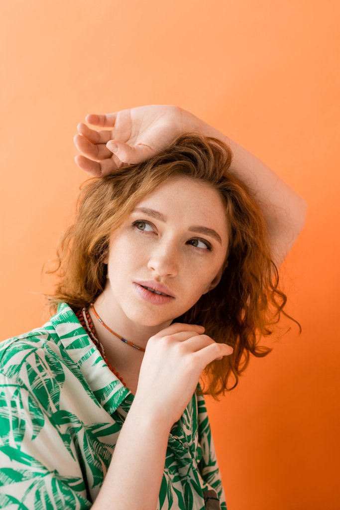 Portrait of dreamy and stylish redhead woman in modern blouse with floral pattern looking away and posing isolated on orange, trendy casual summer outfit concept, Youth Culture - Photo, Image