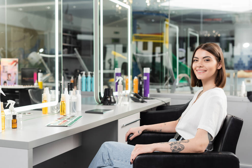 happy salon client, tattooed young woman sitting in chair and looking at camera near hair tools, beauty service, hair extension, hair coloring, hair treatment, blurred background, generation z  - Photo, Image