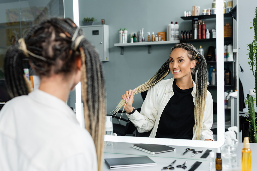 happy client in beauty salon, joyful woman with braids looking at mirror, customer satisfaction, beauty salon, hairstyle, female client with braids,  mirror refection, two ponytails, hair extension  - Photo, Image