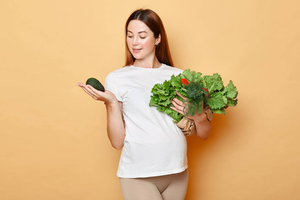Schattige mooie blanke zwangere vrouw met verse groenten poseren geïsoleerd over beige achtergrond kijken naar avocado in haar hand houdt van biologisch voedsel. - Foto, afbeelding