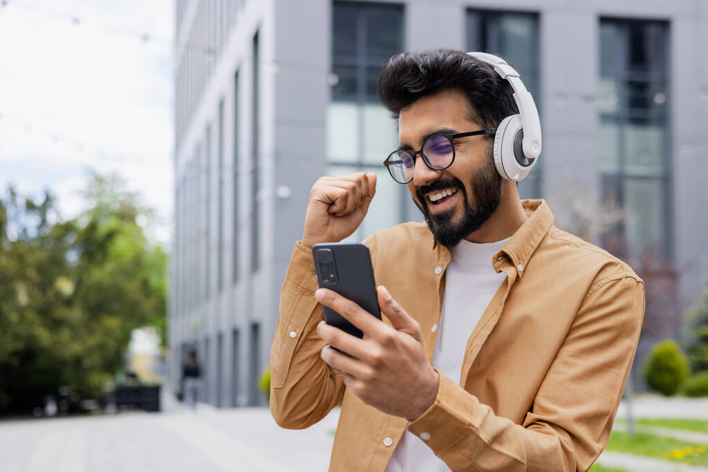 Młody szczęśliwy hindus tańczący na ulicy w słuchawkach, człowiek korzystający z aplikacji online do słuchania muzyki i radia, student w koszuli spacerujący po mieście. - Zdjęcie, obraz
