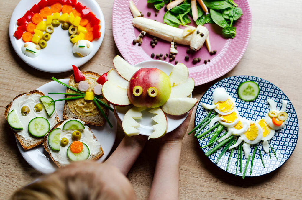 Kädet lapsen lautasella terveellistä hauskaa ruokaa lounaaksi. Apple, muna ja voileipä eri muodossa luovan aamiaisen. Värikäs pöytä lasten ruokaa. Yläkuva. - Valokuva, kuva