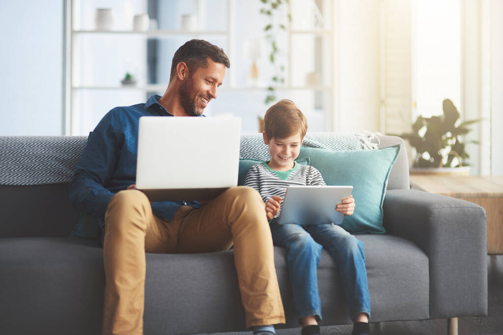 To, nad czym pracujesz synu. wesoły chłopiec i jego ojciec korzystający z laptopa i tabletu cyfrowego podczas siedzenia na kanapie w domu w ciągu dnia - Zdjęcie, obraz