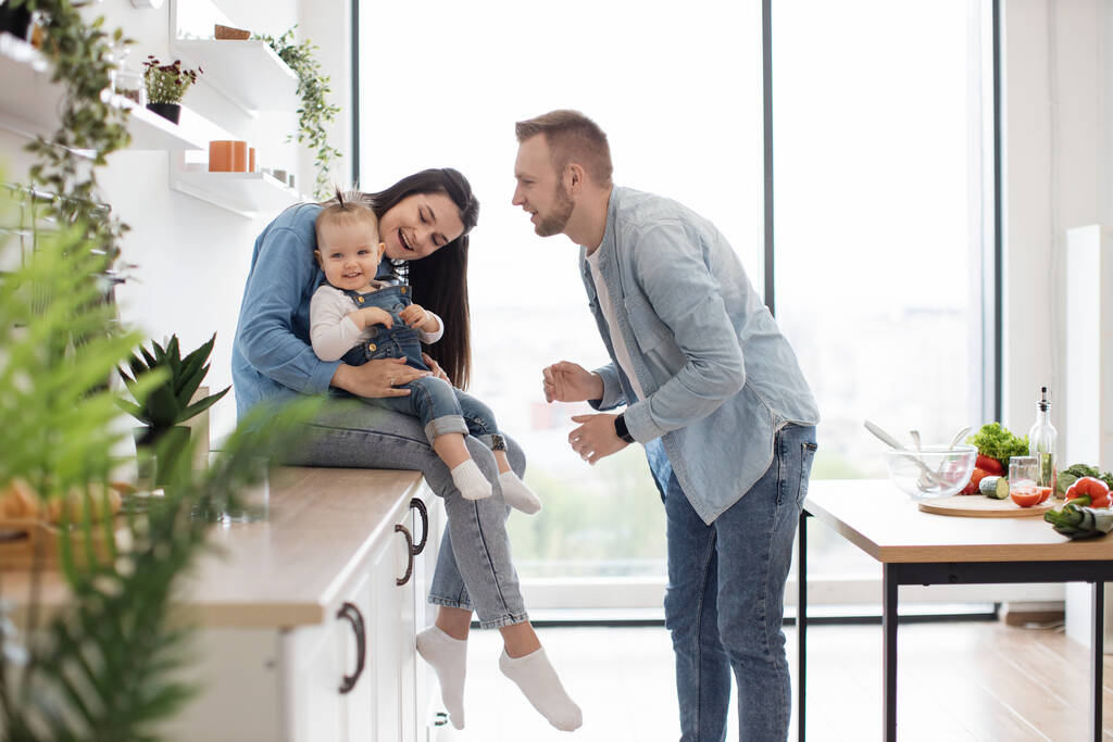 Amusing caucasian man making little girl laugh while standing in open-plan kitchen of modern apartment. Cheerful daughter sitting comfortably on moms knees while having good time with parents. - Photo, Image