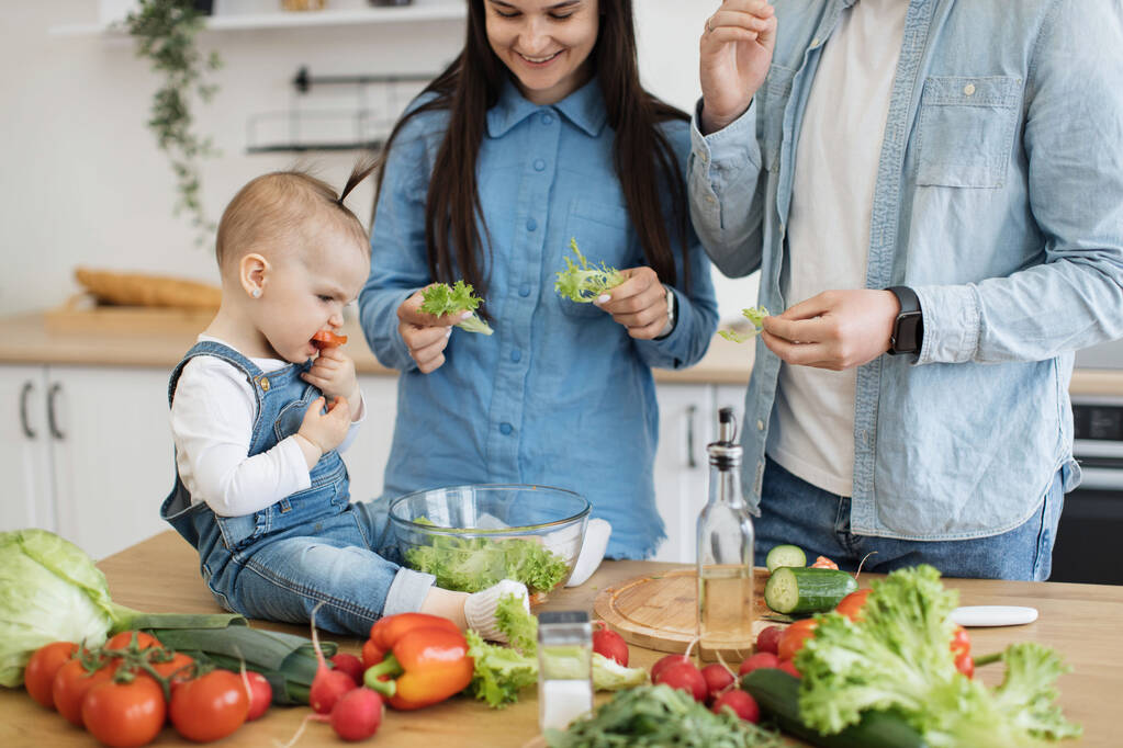 テーブルの上にサラダのボウルの近くにカリカリ野菜を食べる赤ちゃんの娘で笑顔幸せな母親の作物のビュー。新しい食事療法の子供の可能な成分として巻き毛飛び地の新鮮な葉を提供する介護親. - 写真・画像