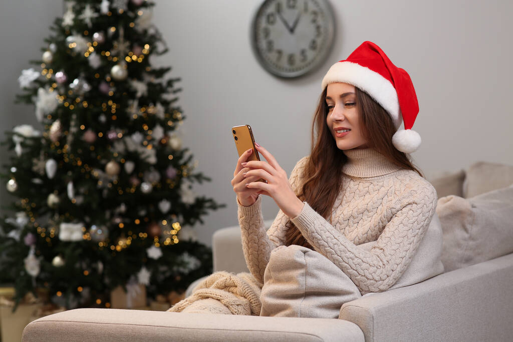 Bonita jovem mulher sorridente em chapéu de Santa usando smartphone perto da árvore de Natal em casa, espaço para texto - Foto, Imagem