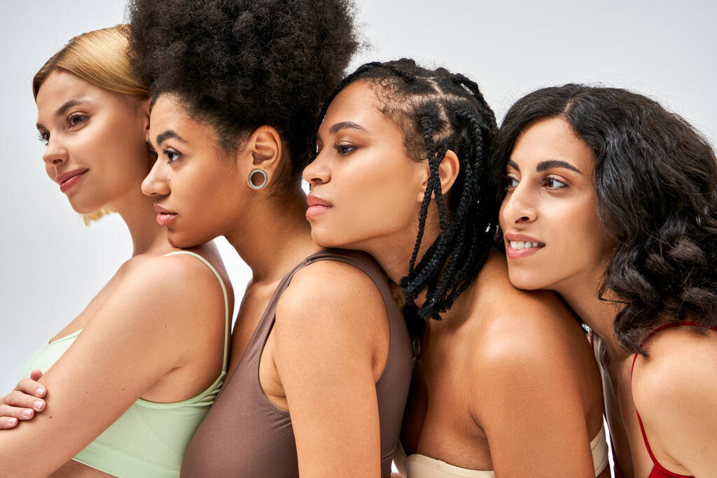 Smiling and multiethnic women in colorful bras looking away while standing next to each other isolated on grey, different body types and self-acceptance concept, multicultural models - Photo, Image