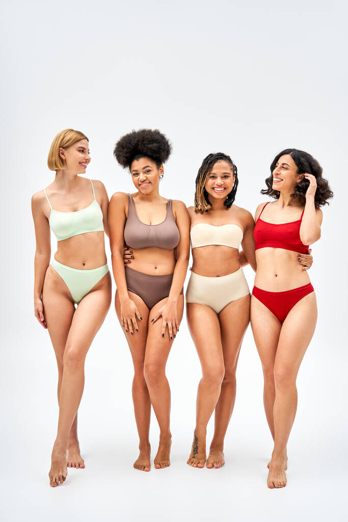 Full length of positive and barefoot multiethnic women in colorful lingerie hugging each other and standing on grey background, different body types and self-acceptance concept, multicultural models - Photo, Image