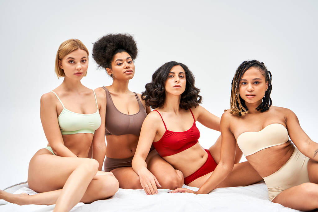 Mulheres multiétnicas em sutiãs coloridos e calcinhas olhando para a câmera juntos enquanto sentado na cama branca isolado em cinza, diferentes tipos de corpo e conceito de auto-aceitação, modelos multiculturais - Foto, Imagem