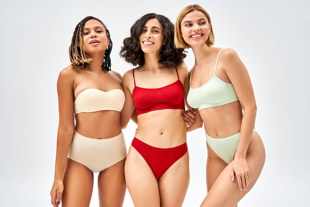 Cheerful young multiethnic women in different colorful lingerie hugging while standing together isolated on grey, different body types and self-acceptance concept, multicultural models - Photo, Image