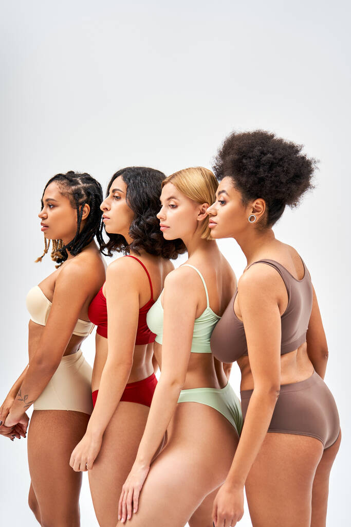 Vista lateral de las mujeres multiétnicas en lencería colorida y moderna mirando hacia otro lado mientras están de pie juntos aislados en gris, diferentes tipos de cuerpo y el concepto de autoaceptación, modelos multiculturales - Foto, Imagen