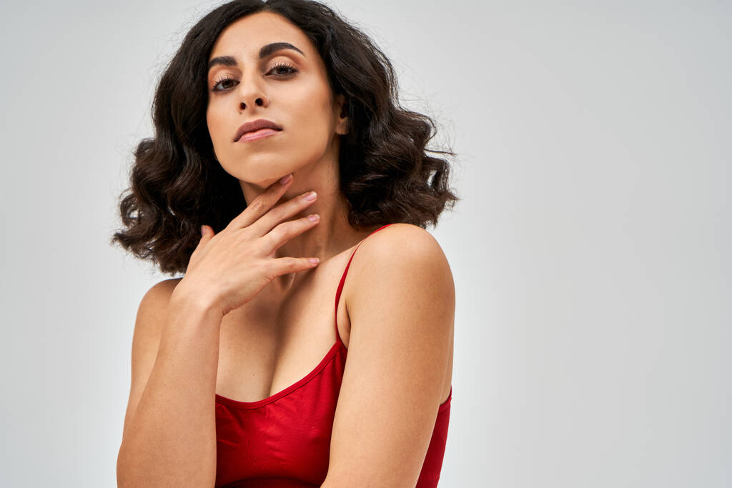 Portrait de femme brune du Moyen-Orient en soutien-gorge rouge tendance touchant le cou et regardant la caméra tout en étant isolé sur le gris, l'acceptation de soi et le concept positif du corps  - Photo, image