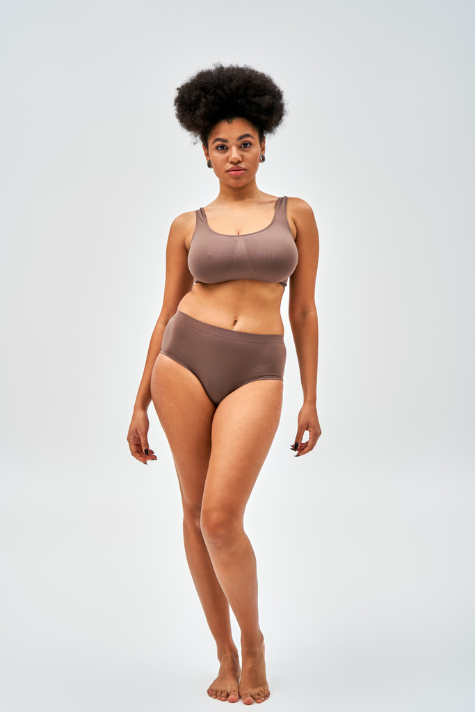 Pleine longueur de femme afro-américaine confiante et pieds nus en lingerie marron regardant la caméra tout en se tenant debout et posant sur fond gris, l'acceptation de soi et le concept positif du corps  - Photo, image