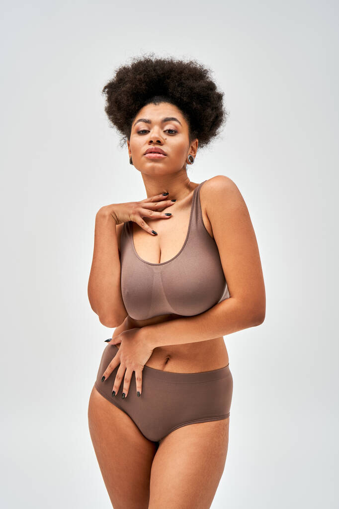 Портрет соблазнительной африканской женщины в современном коричневом нижнем белье, касающемся шеи и смотрящей в камеру, стоящей изолированной на сером, самопринятии и позитивной концепции тела  - Фото, изображение