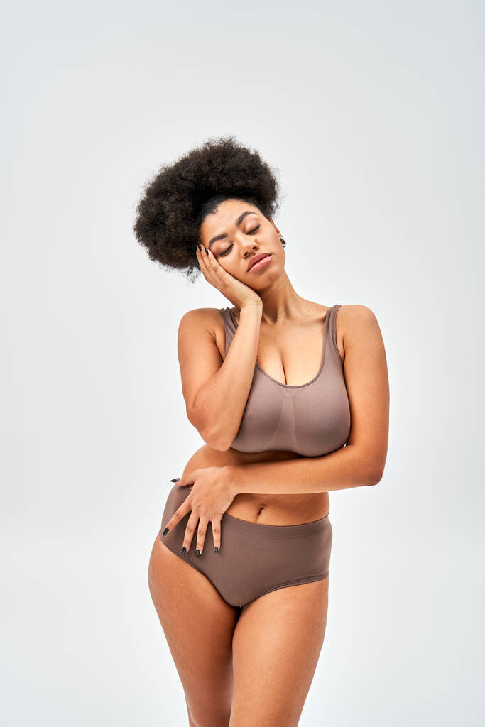 Mujer afroamericana seductora en lencería marrón moderna tocando la mejilla mientras posa con los ojos cerrados y de pie aislado en gris, la auto-aceptación y el concepto de cuerpo positivo  - Foto, imagen