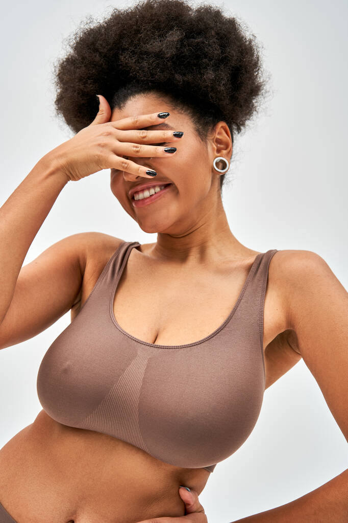 Veselý africký Američan žena v moderní hnědé podprsenka zakrývající tvář s rukou při stání a pózování izolované na šedé, self-přijetí a tělo pozitivní koncept  - Fotografie, Obrázek