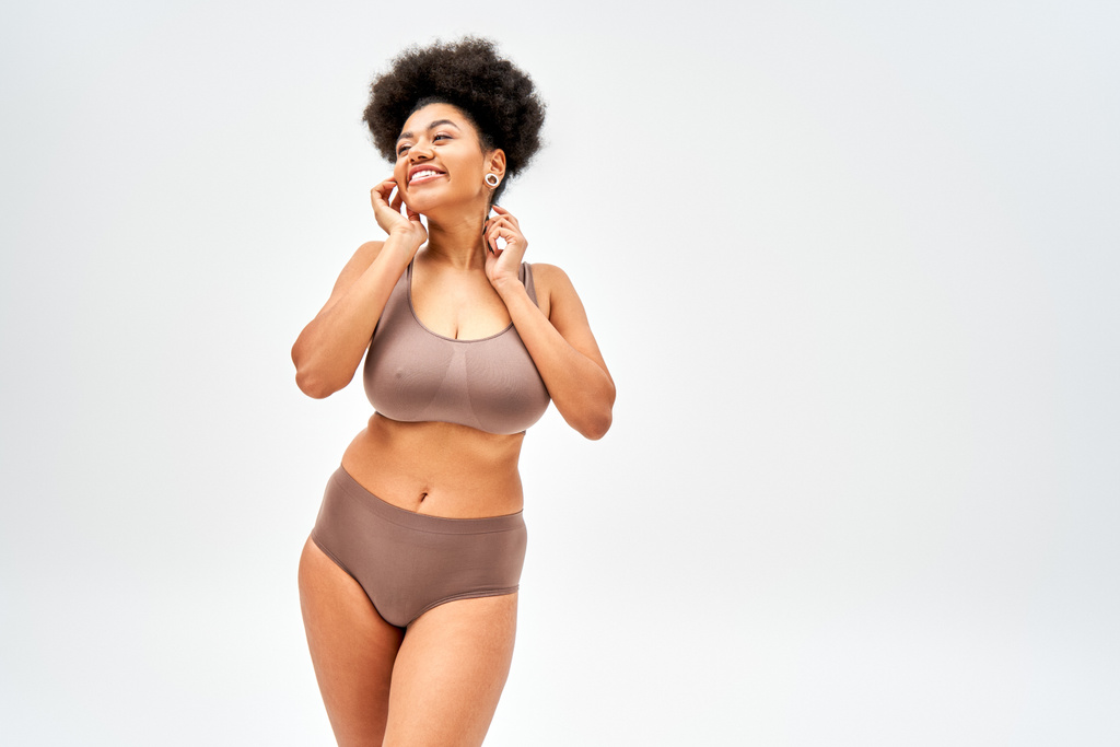 Positive and sexy african american woman in modern brown lingerie touching cheek and looking away while standing and posing isolated on grey, self-acceptance and body positive concept  - Photo, Image